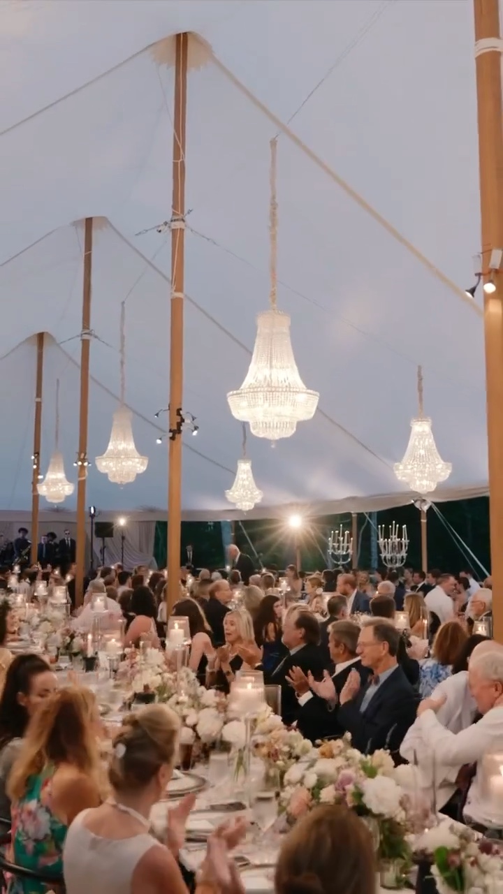 Wedding guests gathered for dinner beneath chandeliers in a waterfront tent